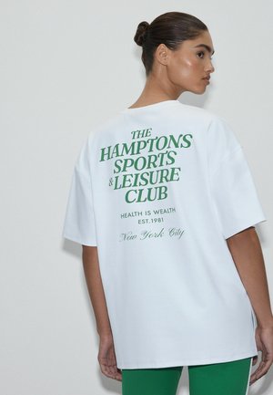 Kobieta z włosami związanymi w kok, ubrana w biały, oversize'owy T-shirt z zielonym napisem "Hamptons Sports & Leisure Club" na plecach, do tego zielone spodnie.