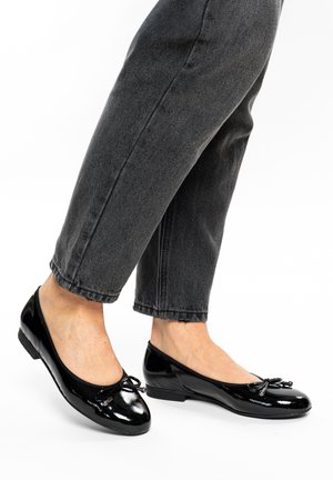 Füße in schwarzen Ballettflats aus lackiertem Leder mit Schleifendetails, kombiniert mit gekürzten dunkelgrauen Jeans, vor weißem Hintergrund.