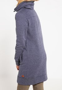 Sudadera larga y ajustada de tejido azul oscuro. Cuenta con un cuello alto, bolsillos laterales y una etiqueta en el dobladillo. Textura suave y diseño minimalista.