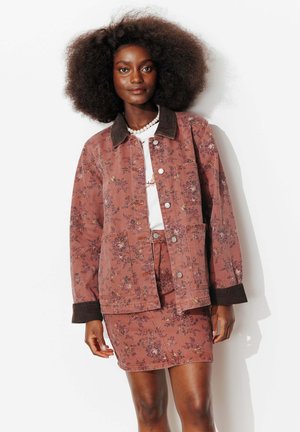 Femme avec une grande coiffure afro portant une veste fleurie couleur rouille et une jupe assortie, une chemise blanche et un collier de perles, debout sur un fond blanc.