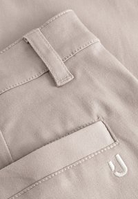 Béžová bavlnená tkanina s hladkou textúrou, so zošitým pútkom na opasok a nášivkovou vreckom. Prítomný je aj malý biely vyšívaný detail 'U'.