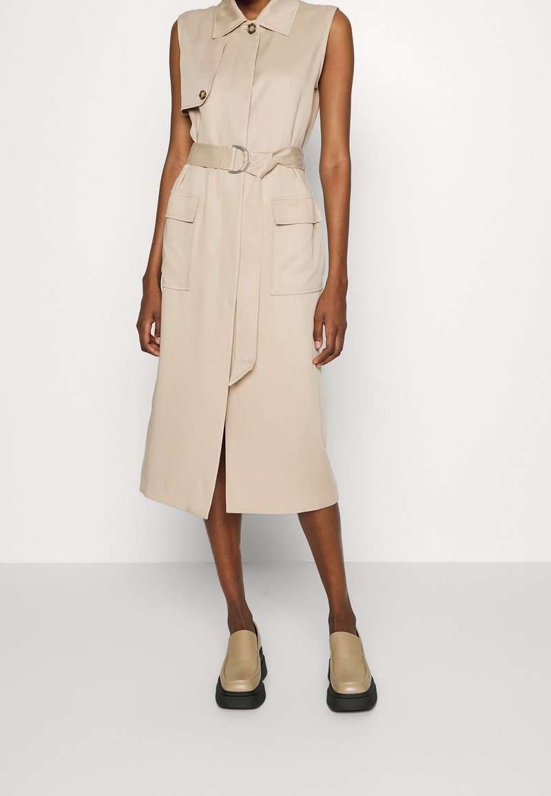 Beżowa sukienka bez rękawów z zapięciem na guziki, paskiem i przednimi kieszeniami; gładka tkanina o długości do połowy łydki. Obuwie z masywną podeszwą.