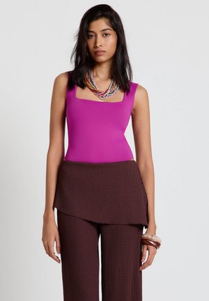 Donna con top senza maniche magenta, pantaloni a vita alta marrone scuro con texture, collana multicolore a più strati con perline e braccialetti spessi.