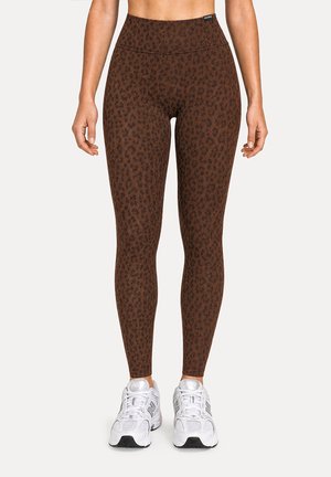 Legging taille haute imprimé léopard marron porté avec des baskets blanches sur une personne debout devant un fond uni.