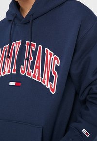 Felpa blu navy con la scritta "TOMMY JEANS" in rilievo rosso. Presenta una tasca frontale, cappuccio con cordino e un piccolo logo multicolore sulla manica.