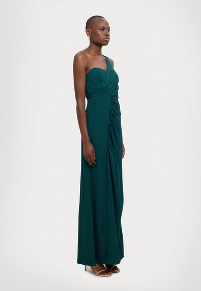 Long, fitted dress in dark green fabric with one shoulder and gathered detailing at the waist. Worn with tan heeled sandals.
