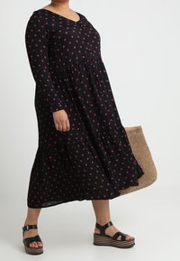 Robe noire à manches longues avec motifs floraux rouges, design à volants, forme évasée, associée à des sandales compensées noires et un cabas en osier.