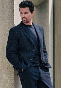 Dunkel strukturierter Blazer mit Revers, getragen über einem schwarzen Rundhals-Shirt, kombiniert mit maßgeschneiderten dunklen Hosen, die sich gegen einen Betonpfeiler lehnt.