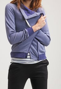 Felpa zip con cappuccio viola e colletto scuro a contrasto, indossata sopra una camicia a righe bianca e blu navy, abbinata a pantaloni neri.