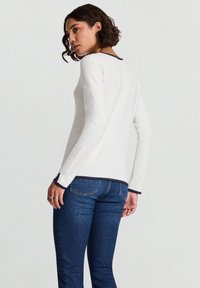 Langärmeliges weißes Oberteil mit dunkelblauem Besatz am Kragen und an den Bündchen, kombiniert mit blauen Jeans aus Denim. Glattes Baumwollmaterial, figurbetontes Design.