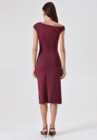 Bordeaux-farbenes, figurbetontes Kleid mit schulterfreiem Ausschnitt, mittellanger Säume, subtiler Rückenreißverschluss und glatter Textur. Akzentuiert mit hohen Schuhen.