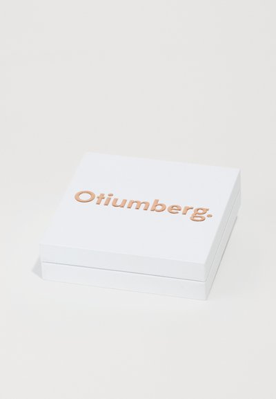 Valge ruudukujuline kast sileda tekstuuriga, millel on tõstetud vaskpruuni värvi logo, millel on kirjutatud "Otiumberg."