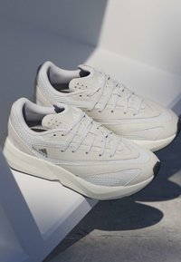 Baskets blanches en mesh et suède avec des semelles épaisses reposant sur une surface blanche inclinée, projetant des ombres sur un sol en béton.