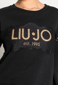 Sudadera negra con un panel de encaje negro texturizado y el logo dorado brillante "LIU•JO est. 1995" en el pecho. Cuello redondo, mangas largas.