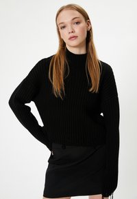 Schwarzer gerippter Strickpullover mit hohem Kragen und überschnittenen Schultern. Ausgefranster Saum und lange Ärmel. Getragen mit einem schwarzen Minirock.