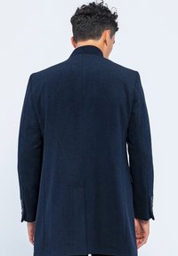 Marineblauwe wolcoat met een spitse kraag, lange mouwen en een getailleerde pasvorm met een rugsplit en knoopdetails op de manchetten.