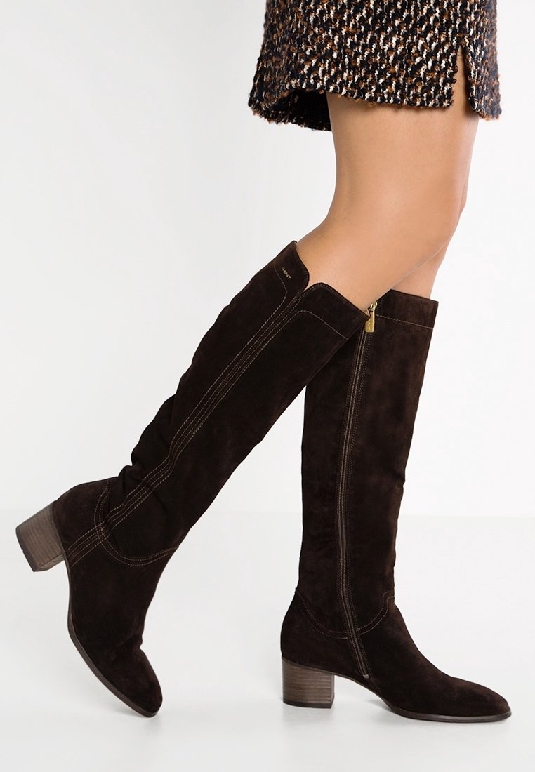 Jambes portant des bottes noires en daim montant jusqu'aux genoux, à talons carrés et fermetures éclair latérales, associées à une jupe à motifs.