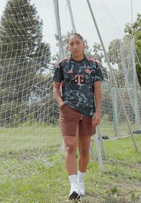 Musta kamuflaažiga spordijerseys punaste aktsentidega, pruunid lühikesed püksid, valged sokid ja mustad spordijalatsid. Taustal jalgpallivärava võrk.