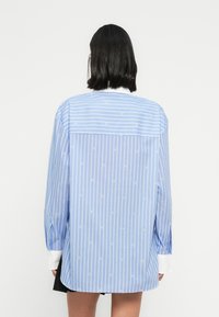 Modrá pruhovaná košeľa s voľným strihom, bielymi manžetami a drobnými vzorovanými detailmi. Dlhé rukávy a zadný diel s preložením dotvárajú dizajn.