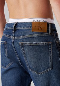 Jeans de denim azul oscuro con una superficie texturizada, que presentan una etiqueta de cuero marrón grabada con "Calvin Klein Jeans" en la parte trasera de la cinturilla.