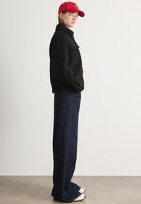 Sort fleecejakke med høj krave, parret med mørkeblå vide jeans og en rød kasket. Skoene er hvide med sølvdetaljer.