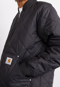 Sort quiltet bomberjakke med ribbede manchetter, lynlås og sidelommer med gul logopatch. Glat stoftekstur.