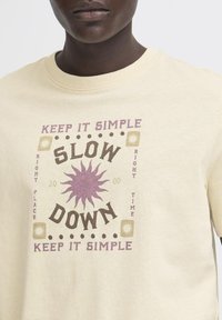 Kremfarget bomulls T-skjorte med grafisk trykk av ordene "SLOW DOWN" i mørkebrunt og lilla, omgitt av solmotiver.
