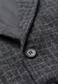 Grijs gemêleerd stof met een ruitpatroon, zwarte knoop met vier gaten, en contrasterende gladde voering. Close-up van de revers.