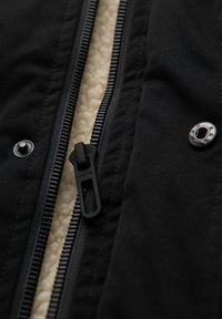 Tejido de chaqueta exterior negra con un suave forro de franela crema. Cuenta con una cremallera negra y herrajes de broche plateados a ambos lados.