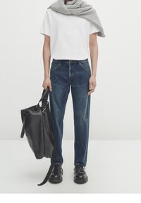 Tummansiniset farkut suoran leikkauksen kanssa, yhdistettynä valkoiseen pyöreäkaulaiseen t-paitaan. Harmaa neulehuivi roikkuu olkapäillä. Mustat nahkakengät.