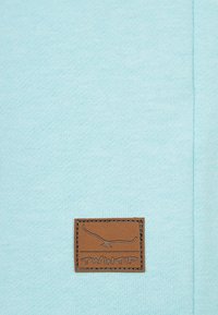 Ljusblått tyg med en liten brun rektangulär läderlapp som visar en stiliserad fågel och texten "TWINTIP" centrerad nära botten.