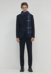 Completo blu navy con una finitura testurizzata, abbinato a una sciarpa a righe blu e grigia. Il look è completato da scarpe nere con lacci.