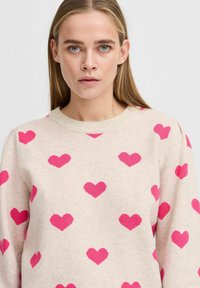 Pull léger beige avec un motif de cœurs roses, texture tricotée, col rond et manches longues. Aucune autre détail de style présent.