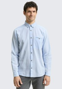 Lichtblauw overhemd met lange mouwen en button-down kraag, subtiele textuur, borstzak en bruine knopenaccenten. Draagt over grijze broek.