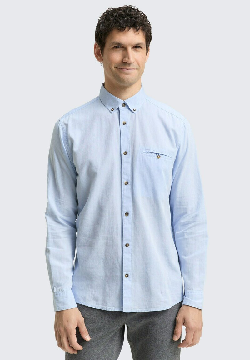 Lichtblauw overhemd met lange mouwen en button-down kraag, subtiele textuur, borstzak en bruine knopenaccenten. Draagt over grijze broek.