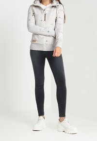 Harmaa vetoketjullinen huppari, jossa on kuviollinen design, nauhat ja tasku, yhdistettynä mustiin skinny-farkkuihin ja valkoisiin suurikokoisiin tennareihin.