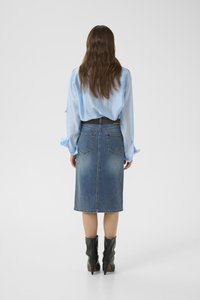 Woman with long brown hair wearing a light blue blouse, knee-length denim skirt, and black heeled ankle boots, standing facing away.