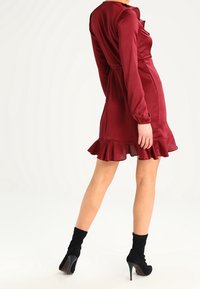 Robe en satin rouge avec des manches longues, ourlet volanté et taille cintrée. Portée avec des chaussettes noires à cheville et des talons hauts. Texture lisse.