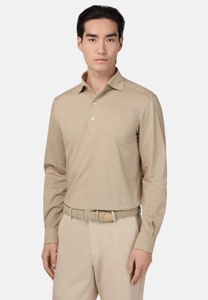 Mand iført en beige langærmet polo-shirt med knapper stoppet ned i lysebeige bukser og et vævet beige bælte, stående mod en ensfarvet baggrund.