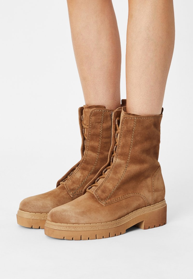 Bottes en daim beige montant à la cheville, avec des lacets à l'avant, des semelles en caoutchouc épaisses et des coutures apparentes, portées sur des jambes nues.