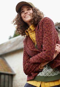 Veste maroon texturée, chemise à carreaux jaunes, pull vert, accents à motifs, et un chapeau pied-de-poule. Cadre en plein air avec lumière naturelle.