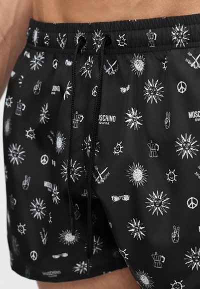 Svarte badeshorts med hvite solansikter, fredssymboler, kaffekanner, solbriller og teksten "Moschino Swim", med svart snor i midjen.