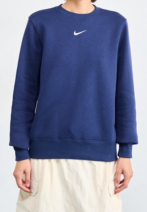 Personne portant un sweat-shirt Nike bleu marine uni à manches longues et une jupe blanche avec des poches.