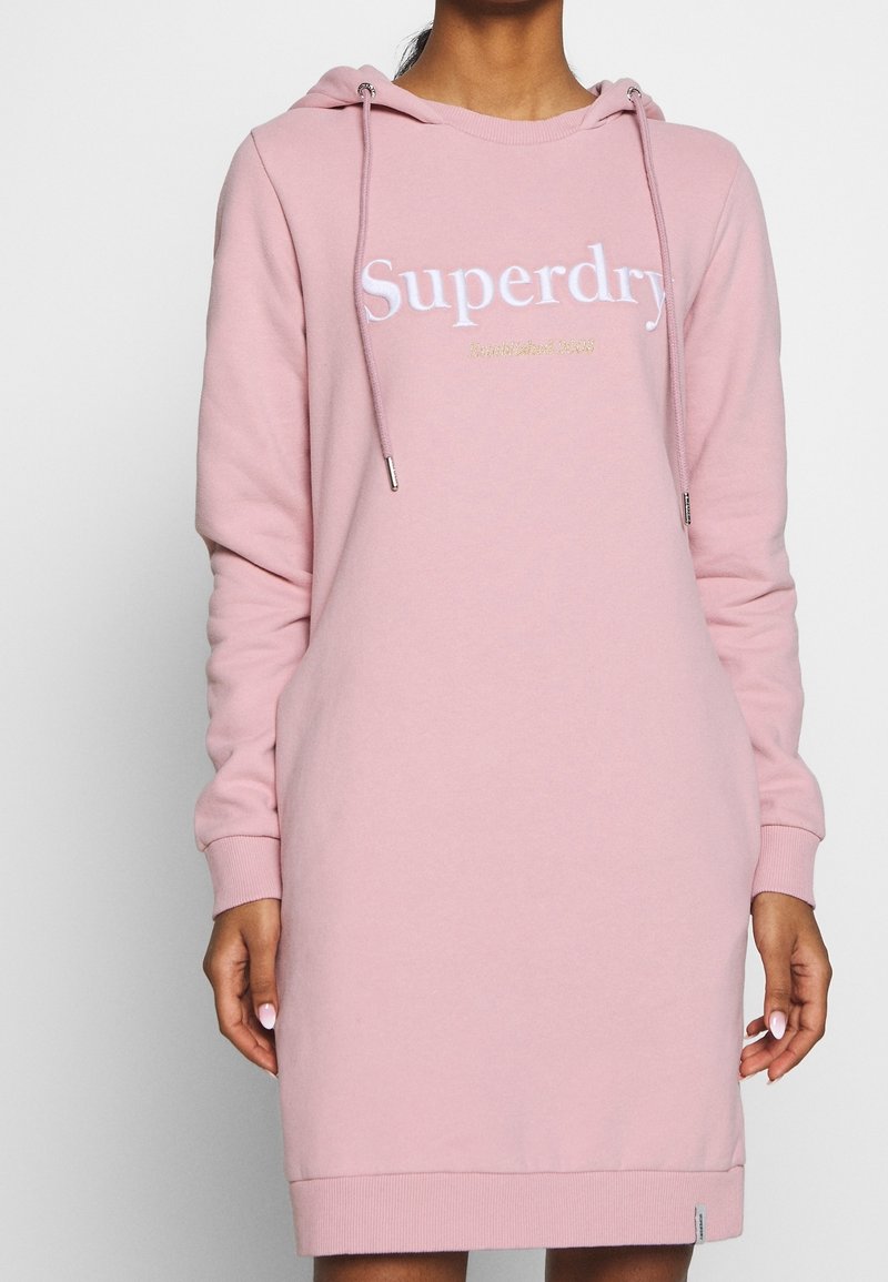 Femme portant une robe à capuche rose à manches longues avec le texte "Superdry Established 2003" sur le devant, debout devant un fond uni.