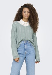Maglione a maglia verde chiaro con motivo a traforo e maniche lunghe, indossato sopra una camicia bianca con colletto, abbinato a jeans blu a vita alta.