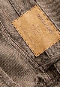 Gros plan sur un tissu denim marron avec une étiquette en cuir fauve cousue indiquant Jack & Jones, Jeans Intelligence, Depuis 1990, Artisanat et Dévouement.