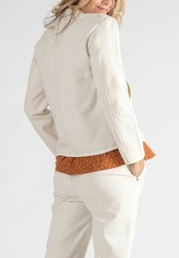 Weiße Denimjacke mit tailliertem Design, ausgestattet mit einer Naht im Rücken und leicht ausgestelltem Ärmel. Kombiniert mit einem orangefarbenen, gemusterten Oberteil darunter.