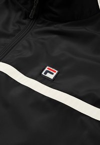 Giacca a vento nera con una striscia bianca e logo ricamato che presenta elementi rossi, bianchi e blu. Materiale liscio e leggero.