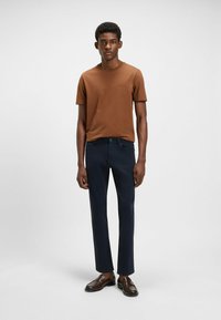 Bruine T-shirt met korte mouwen gecombineerd met marineblauwe broek. Model draagt donkerbruine instappers. Minimalistisch ontwerp met een getailleerde pasvorm.