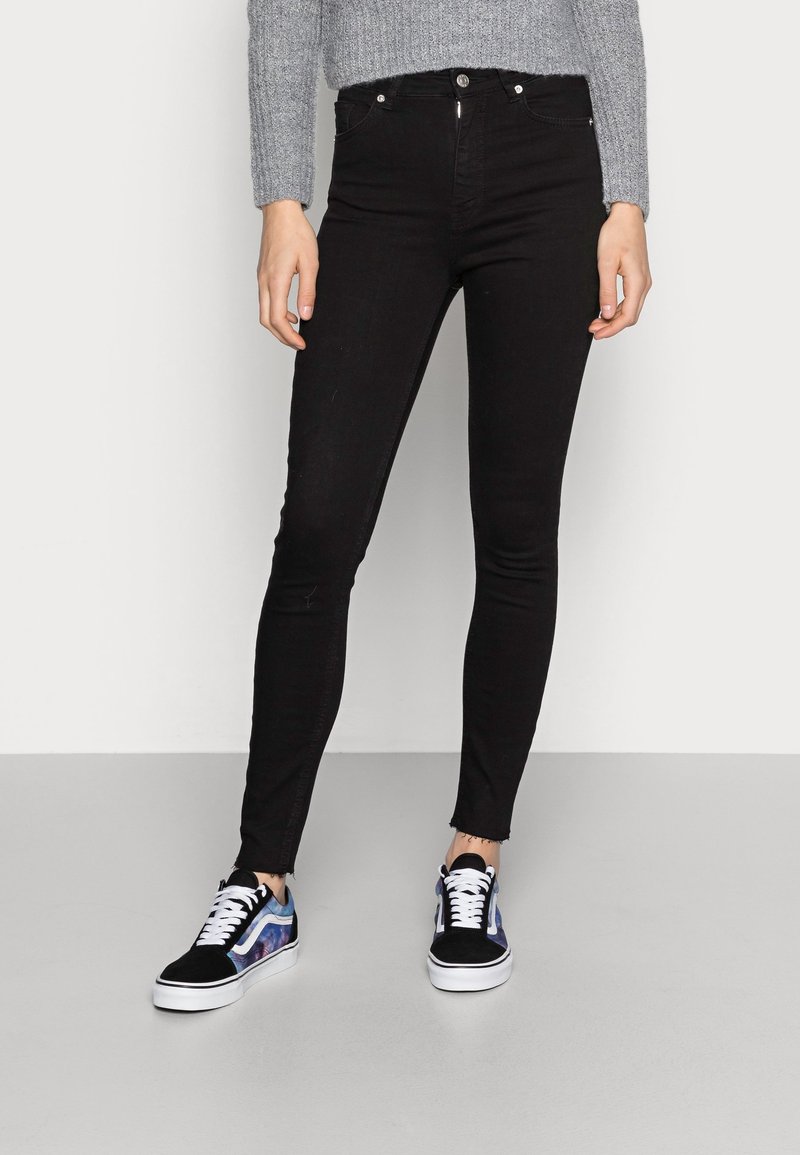 Jeans skinny neri a vita alta con orlo sfilacciato, abbinati a sneakers multicolori con un design nero e blu, su uno sfondo chiaro.